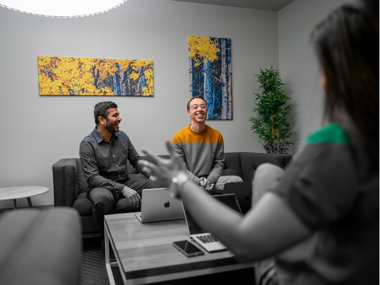 Three AWS colleagues sit together in a shared office space setting. Two men are laughing on a couch sitting across from a woman who is gesturing with her hands. They have open laptops in front of them on a table.